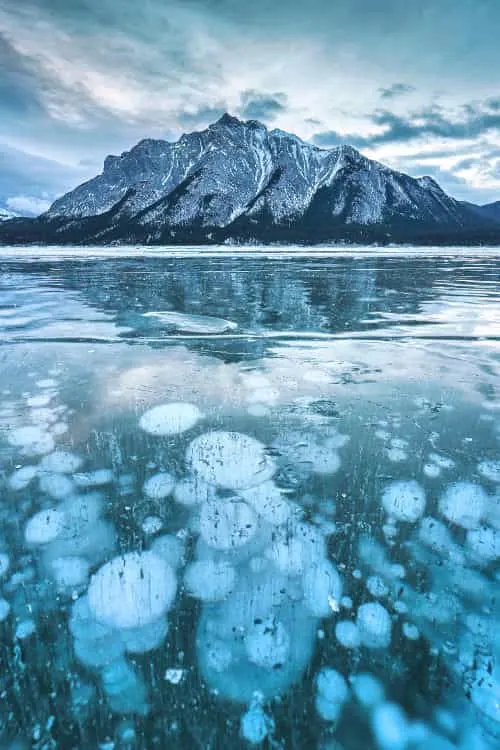 A snowkiter glides across the frozen surface of Abraham Lake, with visible methane bubbles trapped beneath the ice. The shot is taken with a fast shutter speed, freezing the motion of the kiter against the surreal landscape.