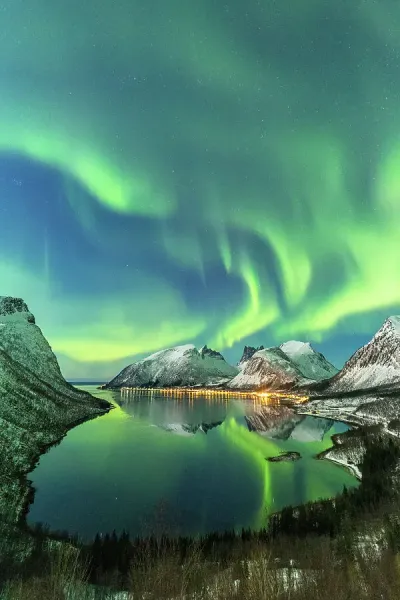 The Northern Lights reflected on the still, dark waters of a fjord near Tromsø, with snow-covered mountains in the background. The long exposure showcases the vibrant colors.