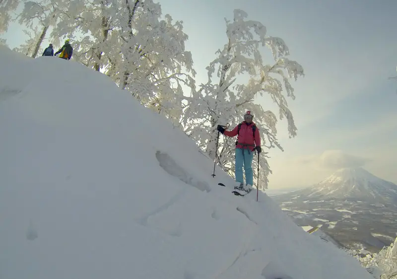 Skiiers carving through deep powder snow on Mount Yotei, with the mountain's snow-covered peak providing a dramatic backdrop.