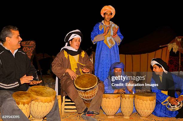 A panoramic shot of the Namib Desert, showcasing the vastness and desolation. A small group of participants are seen navigating the landscape with backpacks, under the guidance of an instructor wearing traditional San Bushmen attire. Focus on the harsh lighting and extreme environmental conditions.