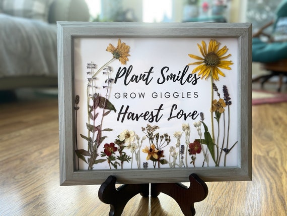 A close-up of pressed flowers (roses, lavender, and calendula) being carefully arranged on acid-free paper using tweezers. The background is a rustic wooden table, emphasizing the natural and handmade aspect of the art.