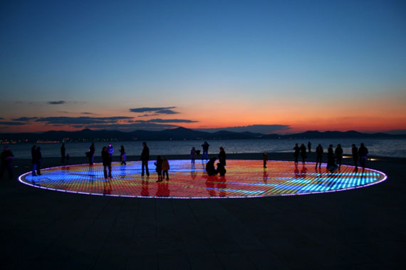 A nighttime view of the Greeting to the Sun in Zadar, Croatia, showing the illuminated solar panels creating a colorful display near the waterfront.