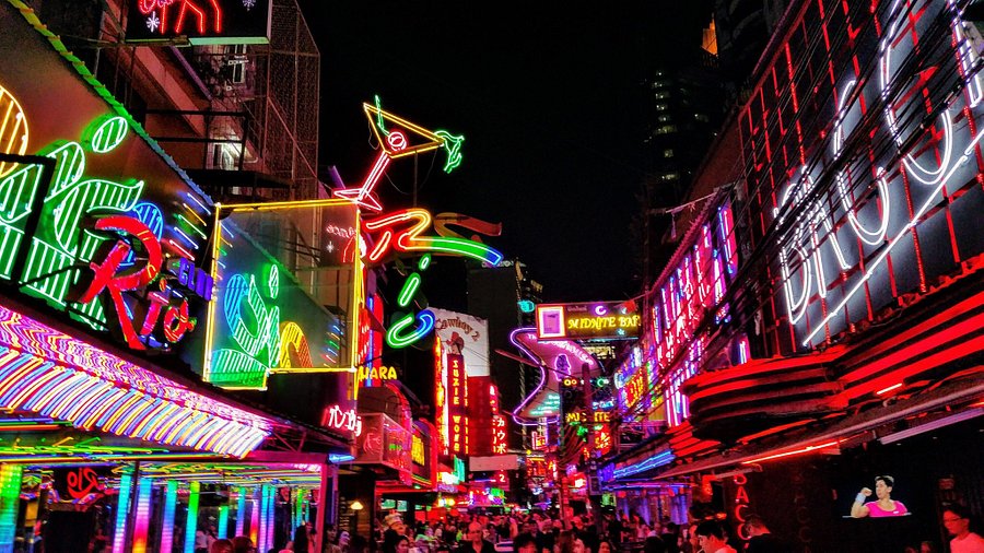 A wide-angle shot of Soi Cowboy at night, capturing the neon signs and bustling crowds, taken with a slightly grainy filter to emphasize the grit. Aim for a shallow depth of field, blurring the background to focus on a specific, interesting detail like a street food vendor grilling skewers.