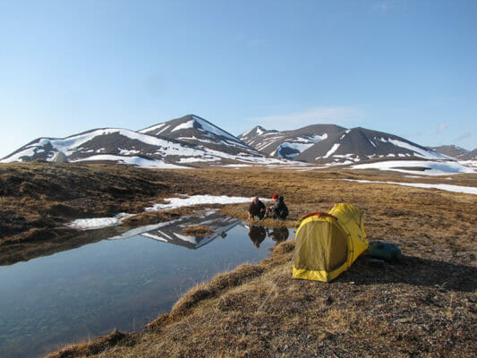 Drone shot capturing the vastness of the Alaskan tundra, with packrafters paddling in the distance. The image highlights the scale of the landscape and the solitude of the experience.
