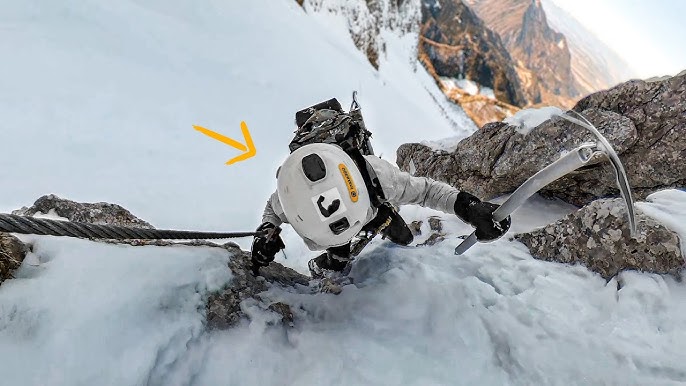 A climber ascending a Via Ferrata route on a snow-covered mountain face. The town of Banff and surrounding mountains are visible in the distance.