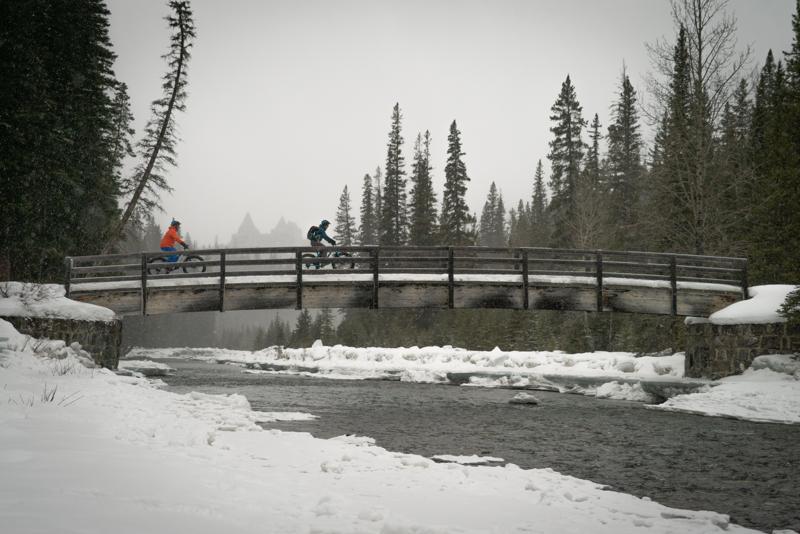 A fat biker rides along a snow-covered path through a dense forest, the Spray River flowing alongside the trail, illustrating the Spray River West Trail's scenic beauty.