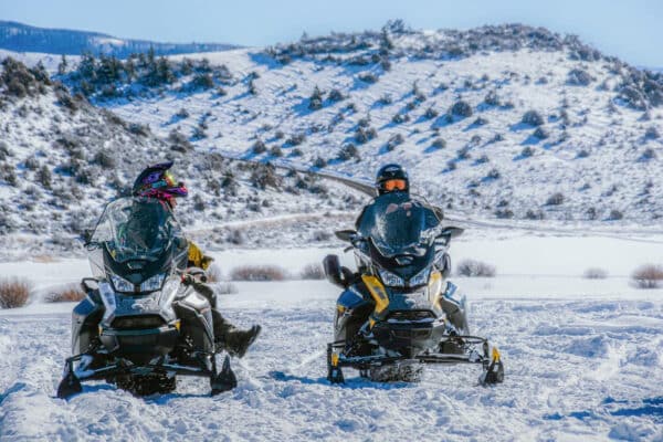 A snowmobile speeds through a snowy landscape with trees and mountains in the background.
