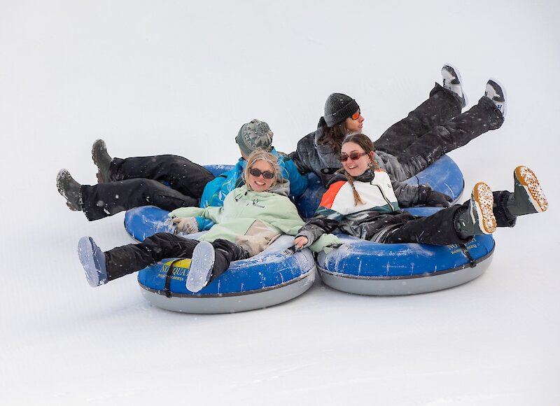A wide shot of people laughing and snow tubing down the runs at Mount Norquay, with the town of Banff and surrounding mountains visible in the background on a sunny day.