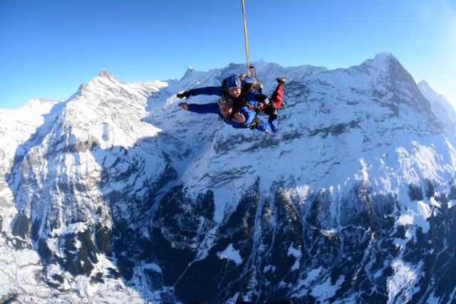 A wide-angle, low-angle shot of a snow skydiving in progress above the Swiss Alps, bright sunlight, blue skies, professional photography, motion blur on the diver, focus on the snowy mountain peaks.