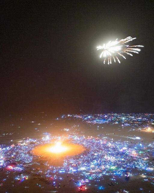 An aerial view captures the vibrant atmosphere of a Black Rock Desert music festival at night, with laser lights painting patterns across the dark sky.