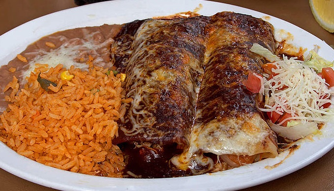 Hotel Emma Library at dusk, lit by vintage lamps, showcasing the floor-to-ceiling bookshelves filled with cookbooks, with a shallow depth of field emphasizing a stack of well-worn cookbooks in the foreground.