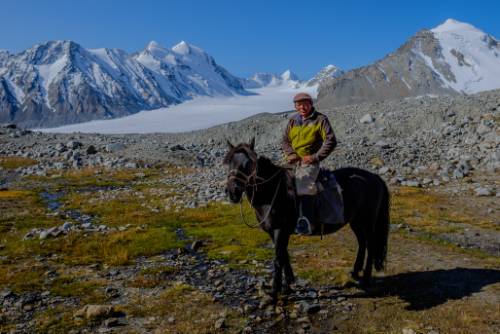 A panoramic view of the vast Mongolian Altai, with a small group on horseback traversing a valley beneath snow-capped mountains.