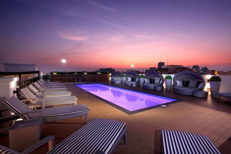 A wide-angle shot of the Khao San Art Hotel rooftop pool at sunset, capturing the backpackers and the city skyline. The shot is slightly overexposed to create a dreamy, warm atmosphere.