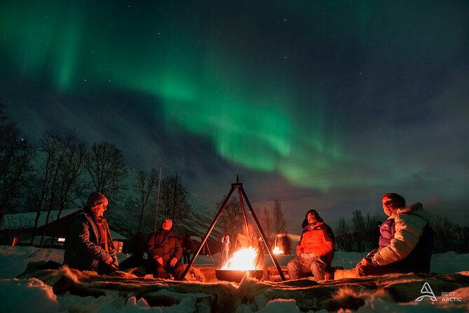A wide-angle shot capturing the Northern Lights dancing above a snow-covered forest, with silhouettes of snowshoers and a Sami guide gathered around a crackling bonfire in the foreground. The aurora should be vibrant and dynamic, showcasing its green and purple hues. Use a slow shutter speed to capture the movement of the lights, creating a sense of awe and wonder.