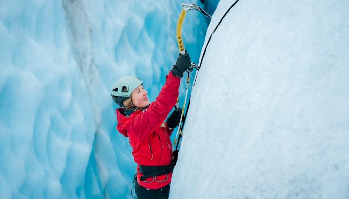 Close-up shot of an ice axe firmly embedded in bright blue ice, showcasing the texture and depth of the glacier ice with a climber in the blurred background