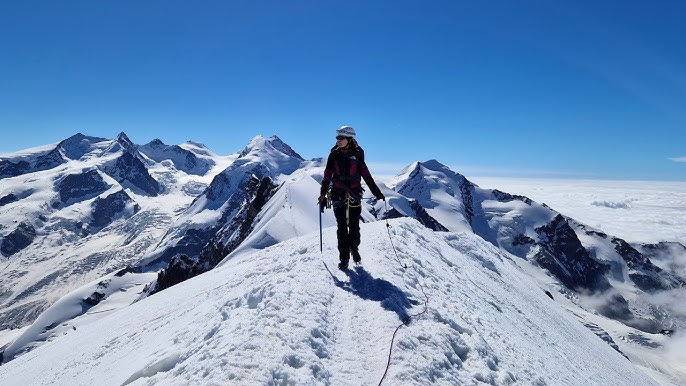 A group of mountaineers roped together, ascending a snow-covered ridge on the Breithorn in the Swiss Alps. Focus on the texture of the snow, the climbers' gear (crampons, ice axes), and the dramatic, jagged peaks surrounding them.