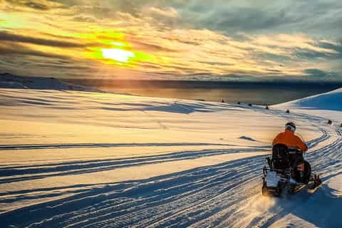 Action shot of a snowmobiler silhouetted against the setting sun on Langjökull Glacier with the vast expanse of snow and ice in the background. A smaller inset photo of the book cover of one of Þórdís Gísladóttir's book, 