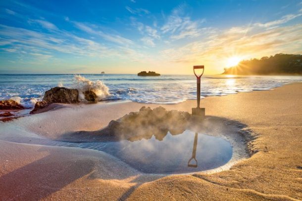 People digging in the sand at Hot Water Beach, creating shallow pools that fill with steaming water. The ocean is visible in the background.