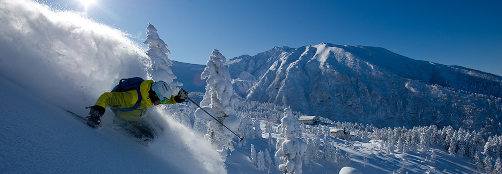A snowboarder carves through pristine powder with a helicopter hovering in the background
