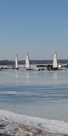A brightly colored ice yacht skimming across a vast, white frozen lake under a clear blue winter sky. High shutter speed to freeze the action and showcase the ice spray.