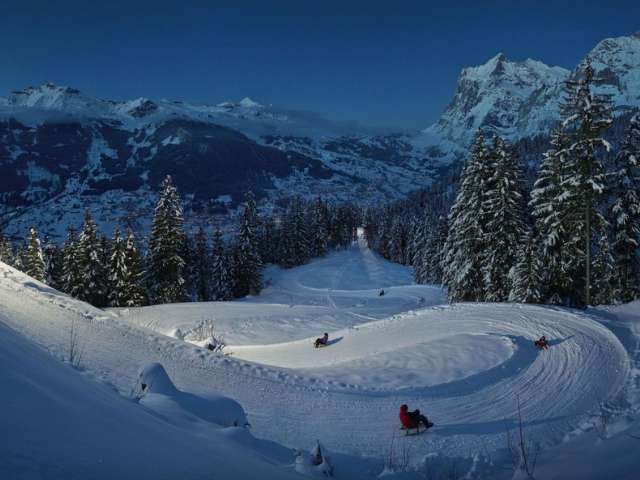 An aerial shot of Ice Magic Interlaken at night, illuminated with colorful lights, showing skaters gliding along the ice. The lights and skaters create a magical atmosphere.