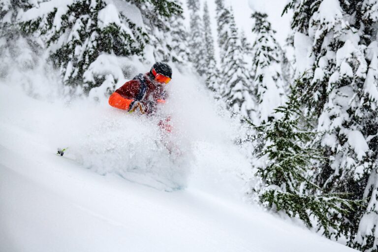 A helicopter landing atop pristine powder in Revelstoke, with a single female skier preparing to descend, her silhouette against the vast white landscape
