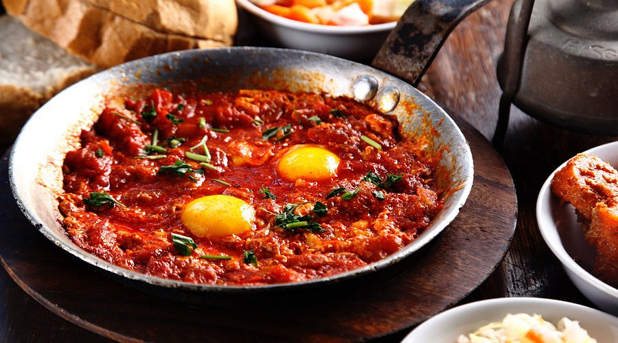 A sizzling pan of shakshuka at Dr. Shakshuka, featuring eggs poached in a spicy tomato sauce, garnished with fresh herbs, served in a rustic pan, and representing the restaurant's lively atmosphere and traditional dish
