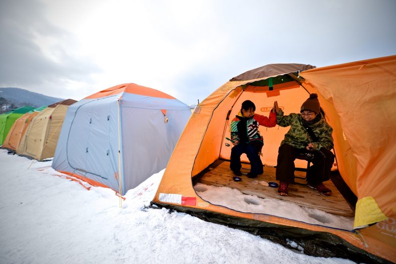 Picture this: Low-angle shot showing a fisherman bundled in warm clothing, illuminated by the soft glow of the interior of his tent, as he pulls up a shimmering wakasagi through the ice.