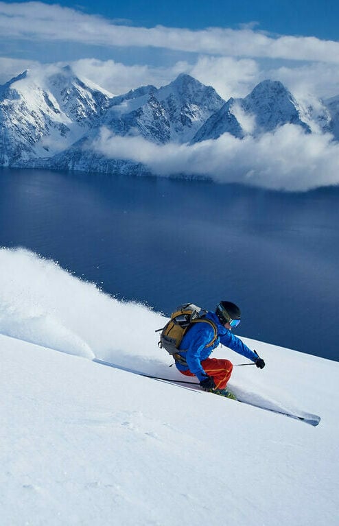 An aerial shot of a lone skier carving down a pristine, snow-covered slope in the Lyngen Alps, with the dark blue waters of the fjord visible far below. The skier should be leaving a clean track in the fresh powder, and the image should convey a sense of freedom and exhilaration. The lighting should highlight the contrast between the bright snow and the dark waters, emphasizing the dramatic landscape.