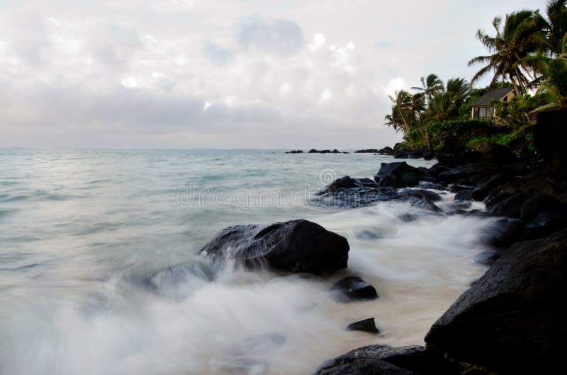 A panoramic shot of the sunrise at Black Rock in the Cook Islands. The sky displays a vibrant spectrum of oranges, reds, pinks, and purples, reflected in the calm lagoon. Soft morning mist adds an ethereal touch to the rocky landscape.