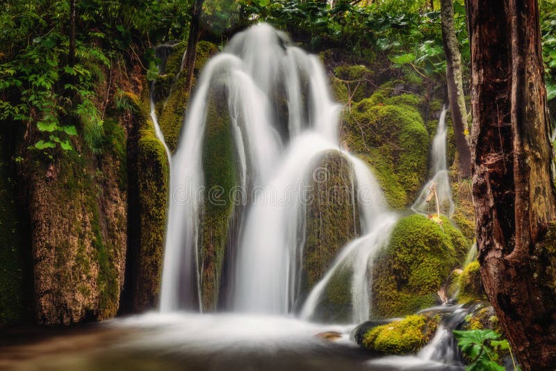 A close-up shot of a series of small waterfalls cascading over travertine barriers, with autumn leaves scattered among the rocks. The water is blurred due to long exposure, creating a silky smooth effect. The lighting emphasizes the interplay of light and shadow on the rocks and water.