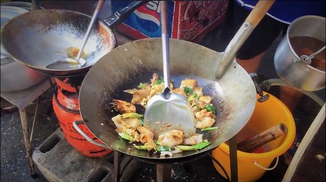 A vendor in Bangkok stir-frying Pad See Ew in a large wok with flames erupting.