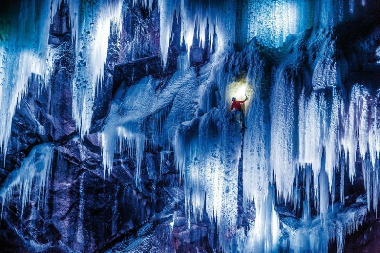 An ice climber ascends a frozen waterfall in the Lofoten Islands, with the village of Svolvær and the surrounding fjords visible in the background.