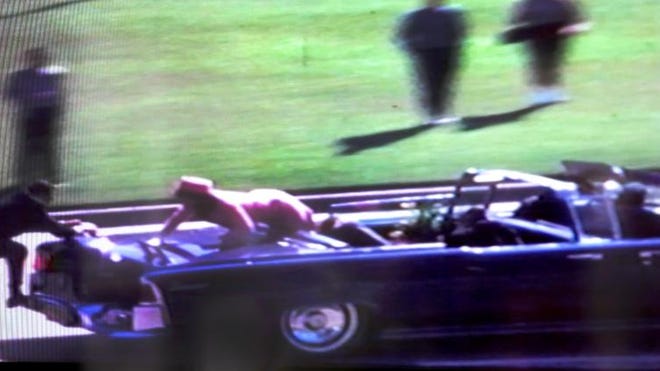A courtroom exhibit featuring a large print of Zapruder film frame 313 displayed on an easel. A pointer rests on the frame, emphasizing the location of the entry wound on Kennedy's head.