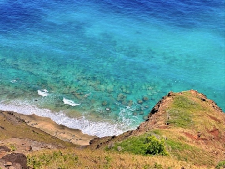 A panoramic shot of Hanakapiai Beach taken from a slightly elevated vantage point along the Kalalau Trail, showing the contrast between the lush green cliffs, the dark sand beach, and the turbulent ocean.