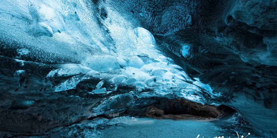 Wide angle shot of a person standing inside a luminous blue ice cave, emphasizing the scale and grandeur of the natural formation