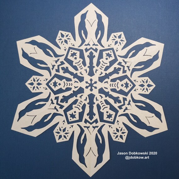 A close-up shot of a hand using Fiskars precision-tip scissors to carefully cut a small triangle along the folded edge of the paper, initiating the snowflake's design.