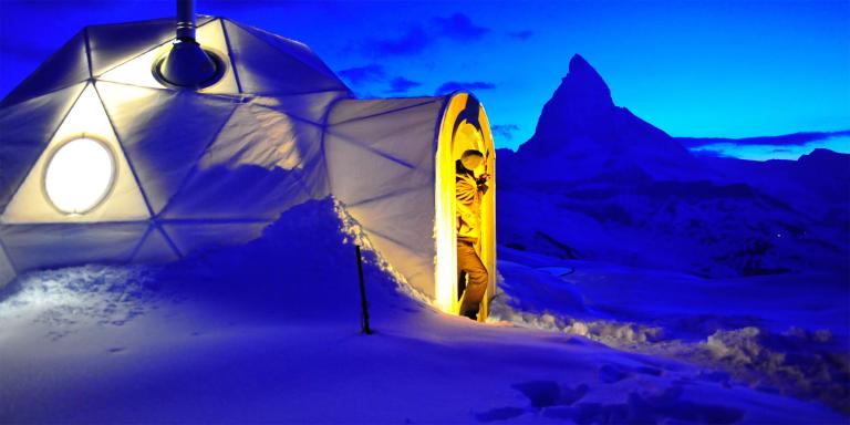 A group of people working together to build an igloo, with the twinkling lights of Grandvalira visible in the distance at dusk. The image captures the sense of teamwork and accomplishment in mastering winter survival skills.