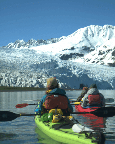 Two kayakers paddle in crystal clear water towards a towering blue glacier, with green mountains in the background.