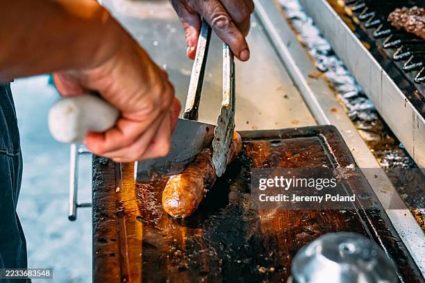 Brightly lit photo of a choripán being prepared on a parrilla, with visible flames and close up on the different condiments being added.