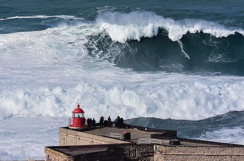 A surfer tackling a massive wave at Praia do Norte, Nazaré, Portugal, with the Nazaré lighthouse in the background, showcasing the scale and drama of big wave surfing.