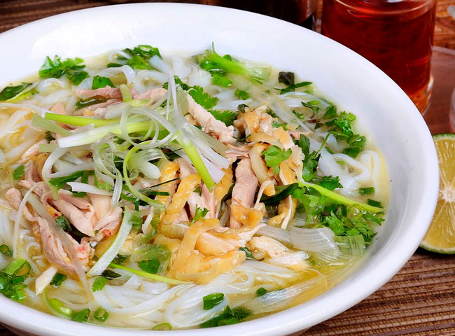 A bowl of Pho, showing the broth, noodles, beef slices, herbs, and lime. The alt text describes the Pho bowl as a rich broth with rice noodles, thinly sliced beef, fresh herbs, and a lime wedge.