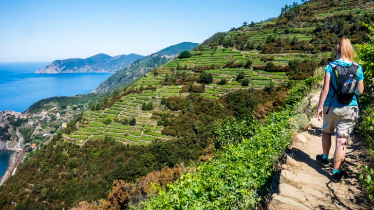 A hiker silhouetted against the blue sea on the Sentiero Azzurro trail in Cinque Terre, with vineyards in the background.