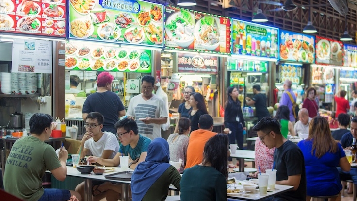A bustling Singaporean hawker center with many food stalls and people eating.
