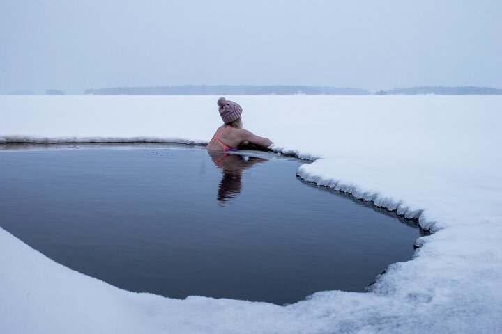 Close-up shot of a person emerging from an ice hole in a frozen lake, steam rising from their body, a traditional Finnish sauna visible in the background, snow-covered trees lining the shore.