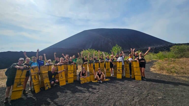 A person volcano boarding down a black volcanic slope, kicking up dust. The sky is bright blue.