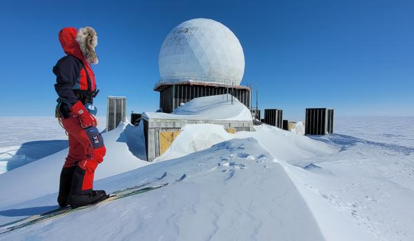 A snowkiter glides across a snow-covered landscape with a large kite overhead. The scene showcases the vastness of the Greenland ice sheet.
