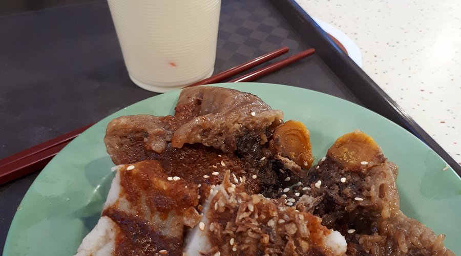 A wide shot of Aunty Kim sitting at a table at Maxwell Food Centre, surrounded by her food. The crowded environment and vibrant atmosphere are captured, showcasing the mix of natural and artificial lighting that accurately represents the conditions