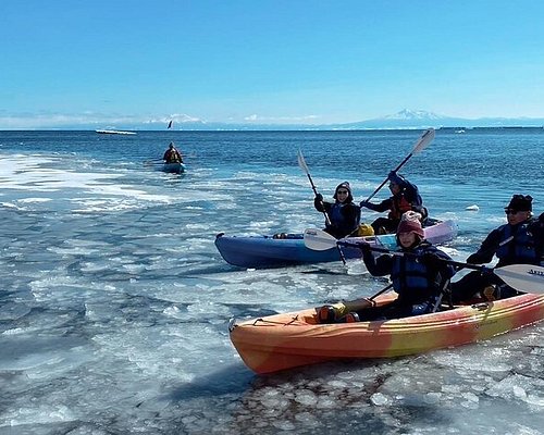 A photo of kayakers in Kokatat dry suits paddling through ice floes in Shiretoko National Park, with a snow-covered cliff face in the background. This demonstrates the adventurous spirit of winter sea kayaking.