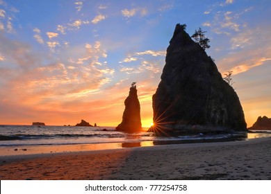 Sea stacks silhouetted against a vibrant sunset at Rialto Beach, with waves crashing on the shore. The sky is filled with orange, pink, and purple hues.